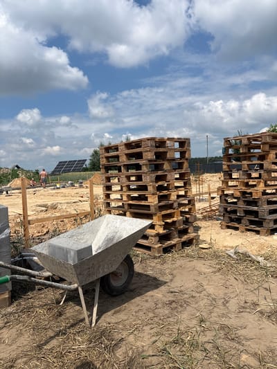 In Opatów, Poland, a construction site is depicted under a partly cloudy sky. In the foreground, a weathered wheelbarrow filled with a concrete block sits on dry, sparse vegetation and dirt. Behind the wheelbarrow, stacks of wooden pallets are piled high. The middle ground reveals the foundation of a building under construction, with disturbed earth and scattered construction materials. Further back, two individuals are present. One person, a bare-chested male, stands near a solar panel array in the distance. Another person is seated. The overall environment is a rural construction zone with elements of both natural landscape and developing infrastructure.