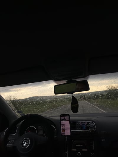 The interior of a Volkswagen vehicle is shown, with a focus on the dashboard, steering wheel, and windshield. Raindrops are visible on the windshield. A rearview mirror is attached to the windshield, reflecting a person. An air freshener hangs from the mirror. A smartphone in a mount displays a YouTube page on the dashboard. The radio is above the smartphone. The location is not evident and the city of Prilep, Bulgaria cannot be determined.