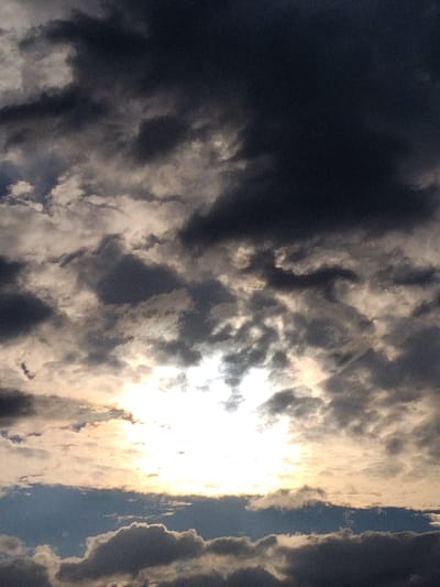 A low-angle shot of a stormy sky is captured, with dark, heavy clouds dominating the upper portion of the frame. The sun, a brilliant white disc, pierces through a gap in the clouds, casting a strong glow and illuminating some of the surrounding cloud formations with a warm, golden light. The lower part of the image shows a clearer patch of sky with a hint of blue, transitioning to more diffuse clouds and a dark cloud bank near the bottom. No people, subjects, or objects are visible. The scene appears to be an outdoor environment, likely captured during daylight hours under threatening weather conditions, possibly in or near Ustyvytsya, Ukraine, given the context provided.