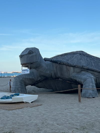 An outdoor daytime shot shows a large, grey, stylized tortoise sculpture on a sandy beach. The sculpture is made of textured material, possibly fabric or canvas, stretched over a frame. Its shell is thatched with straw or similar material. A sign hangs from the tortoise's mouth, reading "BUTAMYATA bay" and featuring a QR code. To the left of the sculpture, a white sofa with decorative blue pillows rests on a woven mat. In the background, there is a body of water, beach umbrellas, and a clear blue sky. The scene appears to be in a resort or beach area.
