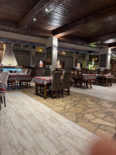 The image depicts the interior of a restaurant in Kumbor, Montenegro. The restaurant features a rustic design with exposed wooden beams on the ceiling and stone walls. Tables are set with patterned tablecloths and chairs. There is a large fireplace on the left side of the image, and decorative tapestries hang on the walls. The lighting appears to be dim, with string lights adding a decorative touch. The floor is a combination of light-colored wood planks and stone tiles. No people are visible in the image.