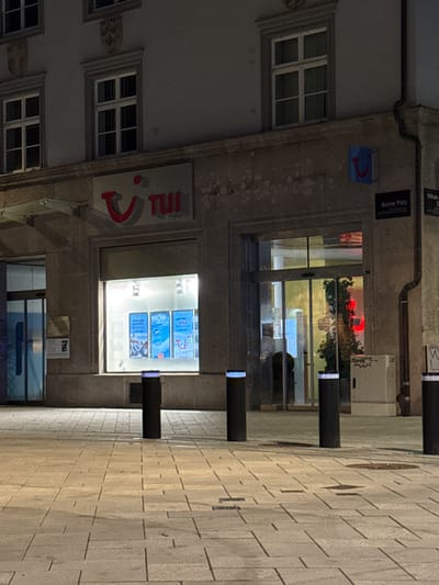 The image captures a view of a TUI travel agency storefront in Innsbruck, Austria, at night. The storefront features a large illuminated sign with the TUI logo, a red smiley face and the word "TUI" in red lettering. Behind the glass windows, promotional posters for travel deals like "WOW Angebote" (WOW offers) and "Last Minute Sale" are visible. The entrance to the store is through automatic glass doors.

Outside the storefront, the pavement is made of light-colored stone tiles, and several black bollards with illuminated tops are placed along the walkway, serving as barriers. In the background, street signs indicate "Bozner Platz" and "Wilhelm Greil Straße." The building itself has a stone facade with architectural details and windows. The lighting suggests it is nighttime, with the storefront being the primary source of illumination. There are no people clearly visible in the image.