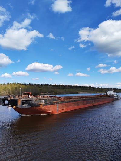 The image shows a barge with the number "N 7903" being towed by a smaller tugboat on a river. The water is dark, and the barge has a red-brown hull with a black upper section.  The sky is blue with scattered white clouds. A forest lines the riverbank in the background. The photograph was taken in Шиндалово, Russia.