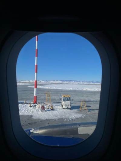 The view from an airplane window reveals a bright blue sky above a snowy landscape. An airport tarmac is visible with a vehicle, likely a pushback tug, and various equipment including yellow stands and a red and white striped pole. A portion of the airplane wing is visible within the window's frame. The clear sky and snow-covered ground suggest a cold climate and sunny weather conditions.