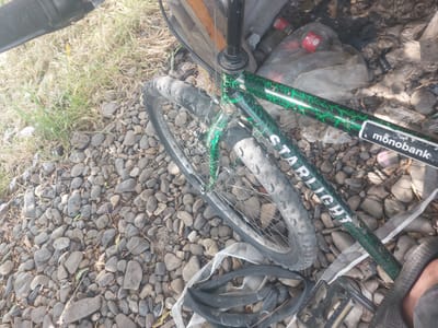 The image shows a close-up view of a BMX bicycle parked on a rocky, unpaved surface, likely a roadside or a natural area. The bicycle has a distinctive green and black crackle pattern on its frame. The visible text on the frame reads "STARLIGHT" and "monobank". The bicycle appears to be in disrepair, with a flat tire and a detached inner tube lying on the ground next to it. In the background, there is a pile of trash, including plastic bags and bottles, some of which appear to be Coca-Cola branded. The lighting suggests it is daytime, with strong sunlight casting shadows. The overall scene evokes a sense of abandonment or neglect, with the bicycle and surrounding debris suggesting a possibly derelict or disused area. The location context of Korolevo, Ukraine, provides a geographical reference for the scene.