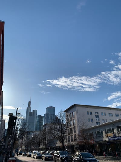 The image shows a street scene in Frankfurt am Main, Germany. The image was captured looking north on a street lined with trees. The street is crowded with cars, some of which are parked along the street. The buildings in the background are high-rise buildings, many of them office buildings. The sky is blue with some white clouds. The location is near the Frankfurt Stock Exchange, the most important stock exchange in Germany, which is located at 50.1111 degrees latitude and 8.6824 degrees longitude. The image is shot from the street level, with a perspective of the city, and is taken in the daytime.