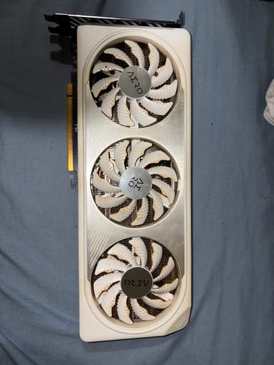 A close-up shot reveals a dirty white graphics card featuring three fans. Each fan has numerous blades with a white and brown color, displaying visible dust accumulation. The central hub of each fan displays the word "AERO." The card's body is white with brushed metal accents, and a metal bracket is visible on the left. Dust and smudges are noticeable across the entire surface of the card.