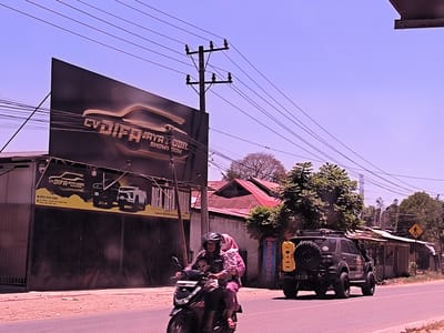 The image depicts a street scene in Keudepanteubreuh, Indonesia. On the left, a building with closed metal doors displays a large sign for "CV. DIFA JAYA MOBIL SHOWROOM." The sign features the silhouettes of automobiles. A utility pole with power lines extends upwards to the right of the building. In the foreground, a person wearing a helmet and floral-patterned clothing drives a motor scooter, carrying another person in pink attire. In the background, a black SUV with a spare tire mounted on the back and a yellow fuel canister is on the right side of the road. A crosswalk sign is visible further down the street, which is lined with structures including homes and shops. The sky is bright and clear.