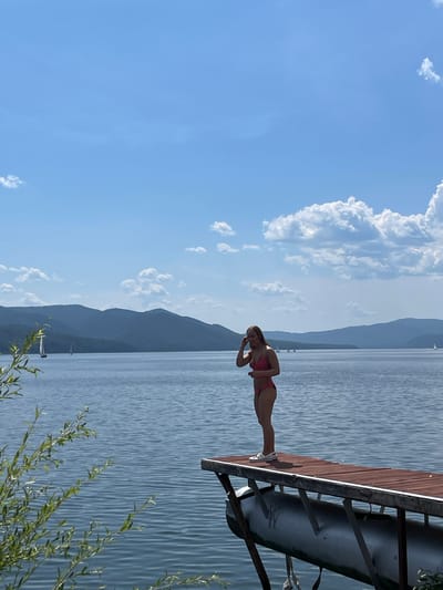 A young woman in a pink bikini stands on a wooden dock on a body of water, with mountains in the background. She is facing to the left, with her right hand touching her hair. The water is blue and calm, with gentle ripples. There are a few sailboats in the distance. The sky is blue with scattered white clouds. The scene appears to be a summer day in a rural or mountainous area. The location is likely near Divnogorsk, Russia, given the mountainous landscape and prominent reservoir.