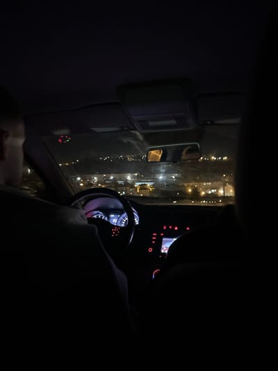 Inside a vehicle at night, a driver holds the steering wheel. The dashboard glows with illuminated gauges and a central display. Through the window, city lights are visible, reflected in the side mirror. The interior is dark, highlighting the illuminated components and external lights. The vehicle is presumably moving, capturing a moment of urban night driving.