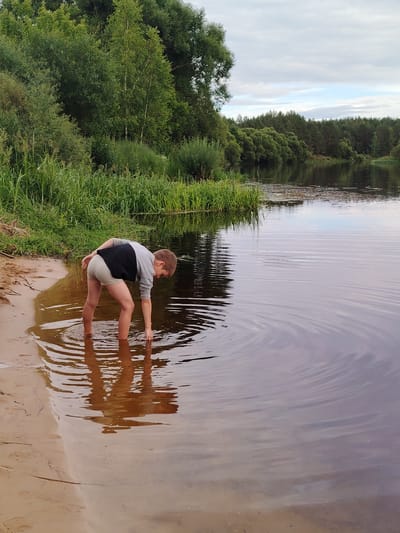 A young boy stands in shallow water at the edge of a river, bending over to touch the water with his right hand. He is wearing a grey and black long-sleeved shirt and light-colored shorts. His legs are submerged up to his knees, and his reflection is visible in the murky water. The riverbank is sandy with some vegetation, and tall green reeds grow further back. Across the river, a dense forest of green trees lines the horizon under a cloudy sky. The scene appears to be set in a rural area, possibly a natural reserve or park. The lighting suggests it is daytime, likely late afternoon or early evening, given the soft light and the overcast conditions. There is no visible text in the image.