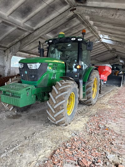 The image portrays an interior view of an agricultural storage structure located in Palamartsa, Bulgaria. A green John Deere 6125 R tractor is the primary subject, positioned in the foreground and angled slightly to the left. It features heavy-duty, deeply-treaded tires with yellow rims, partially covered in dirt. The front of the tractor includes a grill with multiple headlights, a series of green counterweights, and an exhaust stack. A yellow rotating beacon is visible on the roof of its enclosed cab.
Behind the John Deere tractor, further back in the shed, a red agricultural implement, consistent with a fertilizer spreader, is attached to a yellow front-end loader or utility tractor, which is partially visible.
The ceiling of the structure is constructed from pre-stressed concrete panels supported by concrete beams, exhibiting signs of age and dirt accumulation. The interior walls appear to be light-colored, possibly concrete, with visible staining. An open side of the building on the left reveals a blurred, overcast outdoor environment.
The floor is uneven, composed of packed dirt, loose gravel, and a significant concentration of broken red bricks and tile fragments, especially prominent along the right side of the image. Tire tracks are discernible in the dirt beneath the machinery. No individuals are present in the scene.