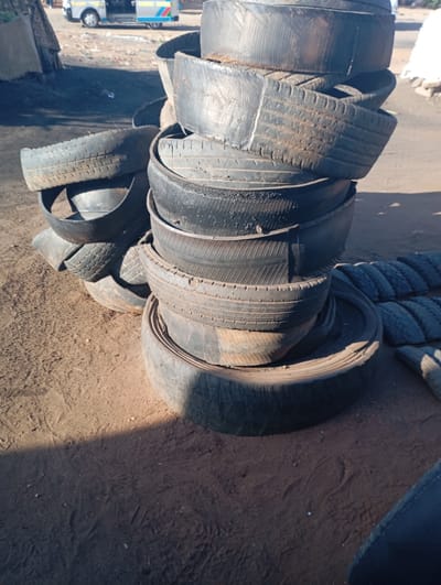 The image captures an outdoor scene featuring numerous stacks of used tires on dry, dusty ground. In the foreground and midground, multiple piles of black tires are arranged, some appearing to be full tires while others are cut into sections. The tires show signs of wear and age, consistent with being discarded or repurposed materials, and are covered in a layer of dust and dirt.
The ground is a light reddish-brown, uneven, and appears parched, suggesting a dry environment. Strong sunlight illuminates parts of the scene, casting distinct shadows across the ground and the tire piles, indicating a clear, sunny day, likely during the mid-morning or late afternoon.
In the background, to the left, a rudimentary structure made of what appears to be corrugated metal or similar material is partially visible. Further back, on the road or path, a white vehicle with prominent blue and yellow stripes can be seen, which resembles a "matatu" (public transport minibus) common in Kenya. The overall setting suggests a commercial area, possibly a tire repair shop, a recycling facility, or a storage yard for used tires, consistent with the location context of Namanga, Kenya. There are no people visible, and no specific activities are taking place beyond the static arrangement of the tires. No discernible text is visible in the image.