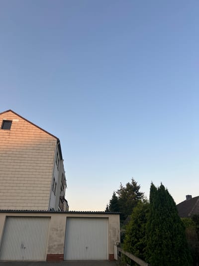 The image depicts a street scene in Schöningen, Germany, under a clear, light blue sky. On the left side of the frame, a multi-story building with a sloping roof covered in light-colored tiles dominates the upper portion. A small, dark rectangular window is visible on the tiled section of the roof. To the right of this building, another taller, multi-story building is partially visible, showing multiple windows and balconies. In the foreground, two white garage doors with vertical paneling are side-by-side, set into a light-colored structure with a corrugated roofline. The garages are positioned on a paved surface. To the right of the garages, lush green evergreen trees of varying heights are densely packed, with a wooden fence partially visible in front of them. In the far right background, a glimpse of a residential building with a dark tiled roof and a chimney can be seen. The lighting suggests it is either early morning or late afternoon, casting a warm glow on the tiled building. There are no people or animals visible in the scene.