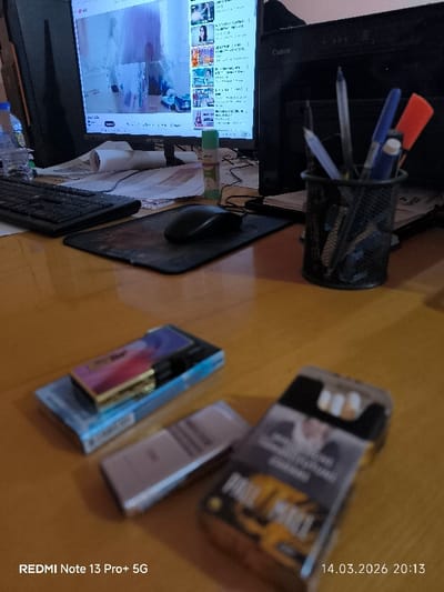 The image displays an indoor desk environment in Pakhtakor, Uzbekistan, captured on March 14, 2026, at 20:13. The central focus is a light-colored, reflective desk surface populated with various items. A black AVTECH computer monitor occupies the left-center background, displaying a YouTube interface with a main video and multiple smaller video thumbnails. To the immediate left of the monitor, a black computer keyboard is positioned, with a black optical mouse on a black mouse pad directly in front of it. A clear plastic water bottle is partially visible on the far left edge of the desk. Clutter behind the keyboard includes several sheets of paper, a rolled white object, and a green and white "Jei-go" glue stick. A black Canon printer is situated to the right of the monitor. A black mesh pen holder containing multiple pens and markers, including blue, black, and red/orange, stands to the right foreground. In the immediate foreground, three packs are visible: a light blue pack, possibly cigarettes, topped with a metallic lighter featuring a pink, purple, and blue gradient design; a silver-grey rectangular pack; and a partially open pack of Pall Mall cigarettes with black and gold packaging, revealing white cigarette filters. No individuals are present in the scene.
