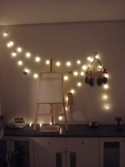The image depicts an indoor scene, possibly a room in Bang Kaeo Subdistrict, Thailand, decorated with string lights. In the center of the frame, an easel stands on a dark countertop, holding a blank canvas. To the left of the easel, a white basket contains several small items. To the right of the easel, there are two small decorative objects and a purple dumbbell. Above the easel, a triangular wall hanging adorned with leaves and branches is suspended. The wall is illuminated by a string of warm white lights that run along the top and down the right side of the wall. The countertop is situated above a set of white cabinets with glass-fronted doors. The overall ambiance is cozy and artistic, with soft lighting creating a warm glow. There are no people present in the image.