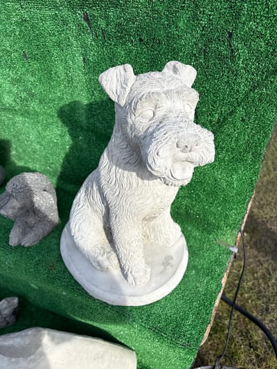 The image depicts a light-colored, detailed concrete statue of a dog lying on its back, paws extended. The statue rests on a patch of artificial green turf. To the lower right of the statue and scattered around it are similarly-colored concrete objects. The surrounding environment, including the city of Chirk, United Kingdom, is not discernible from the image.
