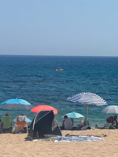 The image shows a sun-drenched beach scene at Platja d'Aro, part of Castell d'Aro i s'Agaró, Spain, with the Mediterranean Sea stretching out to the horizon. In the foreground, several beach umbrellas provide shade: a bright blue umbrella, a red umbrella, a black and white striped umbrella, a teal umbrella, and a light gray umbrella. Beneath these umbrellas, people are relaxing on the sandy shore. Some are seated in beach chairs, while others are lying on towels. A small, dark tent is pitched on the sand, and towels and blankets are spread out. In the middle ground, two individuals are visible in a kayak on the calm blue water. The sky above is a clear, pale blue. The overall impression is one of a leisurely summer day at the beach.