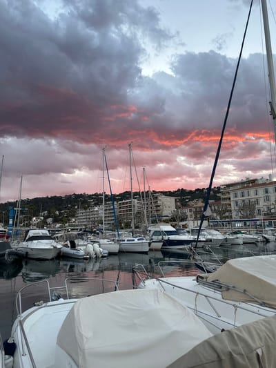 The image captures a view of a marina in Vallauris, France, at sunset. The sky is filled with dark grey and pink clouds. Numerous sailboats are docked in the marina. In the background, buildings and trees line the coast. The image is taken at eye level.