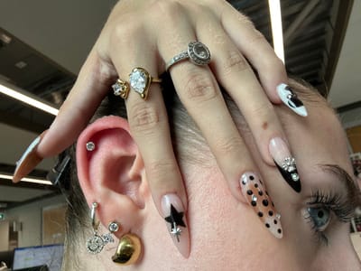 A close-up, eye-level perspective captures the right side of an individual's head and their left hand, situated in an indoor environment in Garston, United Kingdom. The left hand is positioned over the individual's right temple and forehead. The hand displays long, stiletto-shaped artificial nails: the index finger features a black star design with a small diamante on a nude base; the middle finger has black polka dots with small diamantes on a nude base; the ring finger has a black tip with a nude base, adorned with a silver-colored cross-like embellishment and surrounding diamantes; and the pinky finger displays a white tip with a black abstract design and a single diamante. Three rings are worn on the left hand: a silver-colored ring with a central clear stone encircled by smaller stones on the ring finger, and two gold-colored rings on the index finger—one with a large pear-shaped clear stone and another with a smaller round clear stone. A small mole is visible on the back of the hand near the ring finger's knuckle. The individual's right ear is visible with multiple piercings, including a clear stud in the helix, a small clear stud in the lower lobe, a gold-colored dome earring, and a silver-colored hoop earring with a dangling diamante smiley face charm. The right eye, with a blue iris and dark eyelashes, is partially visible in the lower right of the frame. The background is out of focus, revealing fluorescent strip lighting on the ceiling and a faint computer screen on a desk, indicating an office or commercial setting.
