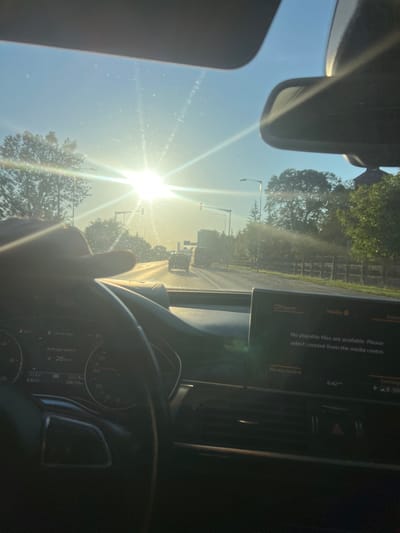 This is a first-person view from inside a car driving on a road. The sun is shining brightly, creating lens flare and glare on the windshield. A hand is visible on the steering wheel. The dashboard displays information including average speed of 26 km/h, an outside temperature of 17.5°C, and the time 6:42 AM. A media screen on the dashboard shows the message "No playable files are available. Please select content from the media centre." The road ahead has other vehicles and trees lining the sides. The environment appears to be a suburban or rural area. The location is identified as Florești, Romania.