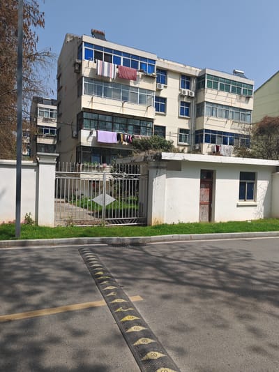 This image captures a daytime exterior scene in a residential area of Jurong, China, under a clear blue sky.

The central subject is a six-story residential apartment building, appearing light beige or pale yellow, with numerous balconies and windows. Many windows feature blue-tinted glass. Multiple external air conditioning units are affixed to the building's facade. Laundry, including towels and various garments, is hanging to dry on balconies and railings at different levels. A solar water heater is visible on the building's flat roofline.

In the foreground, an asphalt road runs horizontally across the frame. A yellow, segmented rubber speed bump with circular symbols imprinted on each segment is prominent on the road surface. A single yellow line marking is present on the asphalt. Tree shadows are cast upon the road. A concrete curb separates the road from a strip of green grass, behind which a white concrete wall forms a boundary.

An ornate silver-colored metal gate, constructed from vertical bars, is integrated into the white wall, providing access to a garden area with green foliage and a visible paved path leading towards the apartment building. A small, white, diamond-shaped sign is attached to the gate.

Adjacent to the gate on the right, a small, single-story white auxiliary building stands, featuring a weathered, reddish-brown wooden door and a single blue-tinted window. Bare tree branches with brown foliage are visible on the far left of the image. No individuals are present in the scene.