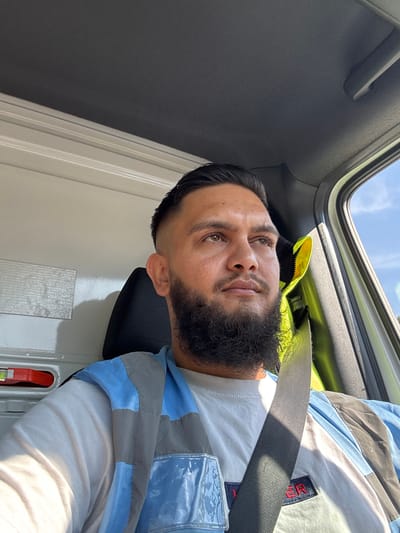 A medium shot captures a man with a full beard, dark hair, and brown eyes from the shoulders up, looking up and to the right from inside a vehicle. He is wearing a light-colored long-sleeved shirt and a high-visibility vest with blue and gray panels. A black seatbelt is diagonally across his chest. The interior of the vehicle is visible, including a black headrest behind him and a white interior panel to his left with a red handle. Sunlight streams in from the right side of the vehicle through a window, illuminating the scene and casting shadows. The background outside the window shows a clear blue sky and some greenery. The man is positioned in the driver's seat, suggesting he is operating the vehicle. The specific location in Namur, Belgium cannot be determined from the visual information provided.