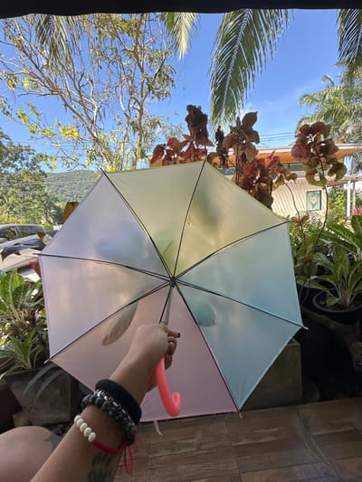 An open umbrella is held against a backdrop of a vibrant outdoor scene. Lush greenery, including palm trees and other plants, frames a clear blue sky. The umbrella's panels are a mix of pale yellow, light blue, and soft pink. A hand is holding the umbrella's pink handle, adorned with bracelets. The image captures a sunny day, with the umbrella providing shade amidst the natural beauty.