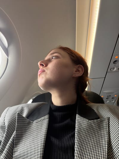 This image depicts an interior scene, understood to be associated with the city of Tuzla, Turkey, possibly during a flight departing from or arriving at the region's airport. A young adult female, with reddish-brown hair styled into a ponytail, is seated within a commercial aircraft cabin. She is viewed in profile, facing left, looking upwards and slightly to the left, with her lips slightly puckered. A small, light-colored stud earring is visible in her right earlobe. She wears a black, ribbed high-neck top beneath a black and white checkered blazer featuring solid black lapels. To her left, a portion of an oval-shaped aircraft window is visible, admitting ambient light. The aircraft's interior consists of light-colored wall and ceiling panels. On the upper right, an overhead passenger service unit is visible, displaying several circular lights, some illuminated amber, and control buttons. A linear strip of overhead cabin lighting runs parallel to the ceiling. A dark-colored seat headrest is partially visible behind the subject.