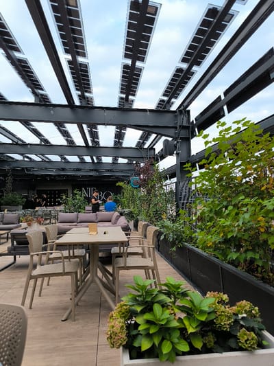 This image captures an outdoor rooftop terrace, identified by the visible "NIEBO" sign as NIEBO Restaurant & Sky Bar in Oława, Poland. The scene is dominated by a large, dark metal pergola structure overhead, which integrates numerous rectangular solar panels arranged in rows. A partially cloudy sky is visible between the pergola elements.
The flooring consists of light-colored tiles or decking. In the foreground and mid-ground, multiple beige/tan chairs with open-weave backs are paired with two-person tables; one table in the immediate foreground features a small menu holder. Further into the background, several grey lounge sofas and low tables are arranged for casual seating.
Extensive greenery is present throughout the terrace. A white planter in the foreground contains lush green plants and light-colored, possibly dried, hydrangea blossoms. Long, dark planters line the right side and mid-ground, holding various bushes and small trees, including a prominent green-leafed shrub.
Four individuals, dressed in dark clothing, are visible in the far background on the left, likely staff members. In the mid-ground, a person in a black shirt stands near a grey sofa, holding what appears to be a menu or tablet, interacting with another individual seated in a blue shirt. String lights are suspended from the pergola structure, and a circular blue light fixture is visible above a planter on the right side.