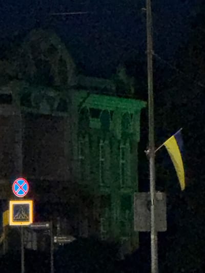 The image shows a Ukrainian flag, with its characteristic blue and yellow colors, flying from a pole. The flag is visible against a dark, night sky. In the background, there is a large, green structure, possibly scaffolding covered by netting, which suggests ongoing construction or renovation. There are also some traffic signs visible in the lower right portion of the image. One sign is circular with a red border and a blue cross, indicating no stopping or parking. Next to it, a square sign with a yellow border contains a symbol that is difficult to discern due to the image quality. The overall scene appears to be an outdoor urban environment at night. The low light conditions and the grainy quality of the image suggest it was taken with a mobile phone camera in a dimly lit area. There are no people visible in the frame. The presence of the Ukrainian flag in this context could signify national pride or support, especially given the ongoing conflict in Ukraine.