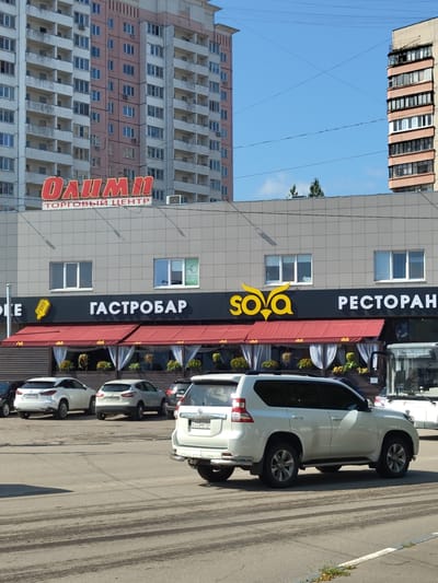 The image captures a street scene in the city of Zhukovsky, Russia, under a clear blue sky. In the foreground, several cars are parked on a paved road with visible tire tracks. A white SUV, a white sedan, and a darker colored SUV are prominent. Behind the cars, a building with a grey tiled facade houses a "GASTROBAR" and a "RESTAURANT," indicated by prominent signage. Above this, a large red sign reads "ОЛИМПИ," with "ТОРГОВЫЙ ЦЕНТР" (Trading Center) below it. The building features several windows, some with closed blinds. In the background, two multi-story apartment buildings are visible. The building on the left is predominantly pink and white, while the one on the right is beige and features balconies and rows of windows. Power lines are visible strung across the sky. The overall impression is of a commercial and residential area on a bright day.