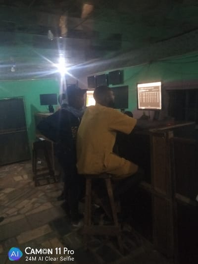 Inside a dimly lit room with green walls, two individuals are present. One sits on a stool, wearing a yellow shirt and focused on a computer screen displaying a grid of data. Another stands to the left, donning a dark jacket and a light-colored neck covering. Additional computer monitors are arranged in the background. A large speaker cabinet is also visible. The overall atmosphere is reminiscent of a betting shop or internet cafe.