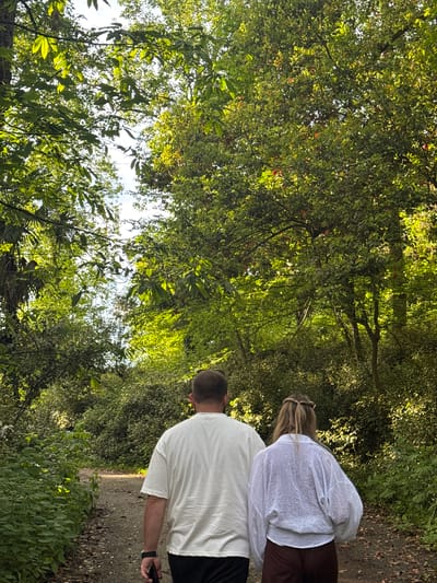 A man and a woman walk along a path through a dense forest in Tsanbaikwara, Abkhazia. The man, wearing a white t-shirt and black shorts, holds a black smartphone in his hand. The woman, ahead of him, wears a white blouse and brown pants. The path is lined with green vegetation and overhead trees. Sunlight filters through the dense canopy.
