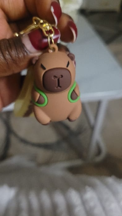 A close-up image depicts a person's dark-skinned hand, featuring dark red manicured fingernails with white accents, holding a capybara-shaped keychain. The keychain, made of light brown rubber or silicone, displays a darker brown snout and ears, and two bright green U-shaped accents on its chest. It is suspended from a gold-colored chain and lobster clasp, with a partially visible translucent yellow rectangular object attached behind it. The hand holds the keychain prominently in the foreground. The background is out of focus, revealing a blurry light-colored, possibly fuzzy, surface in the lower portion and indistinct light grey and white elements, including what appears to be a table surface or furniture. The specific location in Nanyuki, Kenya, cannot be confirmed from the visual information provided in the image.