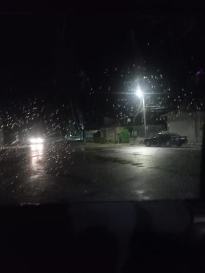 The image captures a nighttime scene on a street in the city of Gradina, Bulgaria. It appears to be raining, as evidenced by water droplets on what seems to be a window or windshield in the foreground, obstructing the view. On the right side of the street, a dark-colored sedan is parked. In the background, streetlights illuminate the wet asphalt, and there are buildings visible. The overall lighting is dim, typical of a nocturnal urban environment during precipitation.