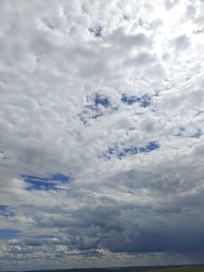The image shows a wide, panoramic view of a cloudy sky over a rural landscape. The sky is filled with a variety of clouds, ranging from thin, wispy cirrus clouds to thicker, darker cumulonimbus clouds. Patches of blue sky are visible between the clouds, indicating a partly cloudy day. The sunlight is diffused by the clouds, creating a soft, even light. The landscape below is a mix of green fields and distant trees, with a hint of what appears to be a small village or scattered houses in the far distance. The scene is static, with no people or animals visible. The overall impression is one of a natural landscape under a dynamic sky, likely in a rural area of Colonița, Moldova, given the context.
