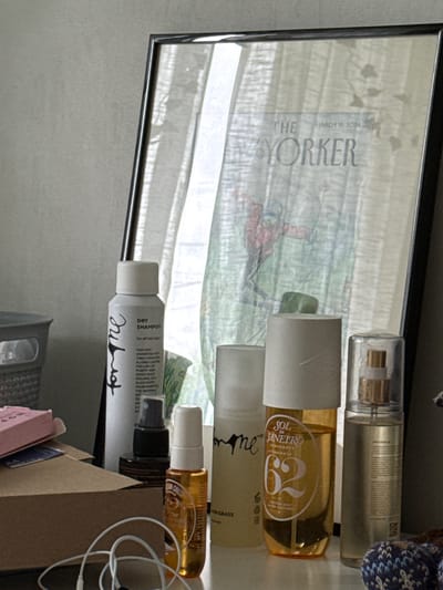 The image depicts a close-up, eye-level view of a shelf with various cosmetic and personal care items arranged in front of a framed mirror. The mirror reflects a blurred image of a window with sheer curtains and a poster or magazine cover that appears to be from "The New Yorker," dated March 18, 2024.
On the shelf, from left to right, are:
* A gray woven basket, partially visible on the far left.
* A small pink box.
* A brown cardboard box.
* A white aerosol can labeled "for me DRY SHAMPOO."
* A small dark bottle with a black spray nozzle.
* A tall, slender clear bottle with an orange liquid and a white cap, labeled "SOL DE JANEIRO."
* A clear spray bottle with a gold pump and a clear cap, containing a light-colored liquid.
* A small glass bottle with an orange liquid and a white pump, labeled "VANILLA CRUSH."
In the foreground, a white pair of earbuds with a wire is coiled on the shelf. The overall lighting suggests an indoor setting, possibly a bathroom or vanity area. The scene is static, with no people present. The location of Коробицыно, Russia, is not determinable from the visual information provided in the image.