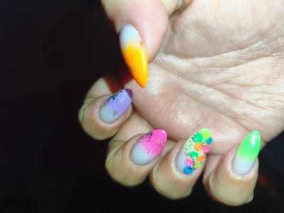 The image shows a close-up of a person's hand with intricately designed fingernails. The nails are long and almond-shaped, featuring a gradient of colors and embellishments. Starting from the thumb (partially visible), the nails display a range of colors including white, orange, purple, pink, yellow, and green. Some nails have glitter or foil accents, while others are adorned with small, colorful flower-shaped studs. The background is dark and out of focus, highlighting the detailed nail art. The hand appears to be resting against the palm of the other hand. There is no discernible information in the image that relates to the city of Komotini, Greece.