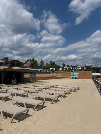 An outdoor leisure area with a sandy surface is depicted under a partly cloudy sky in Hlibivka, Ukraine. In the foreground and midground, numerous unoccupied, light-colored lounge chairs are arranged in orderly rows on light brown sand. To the left, a covered structure, possibly a beach hut, features a brown roof and dark fabric screens, with two white hanging chairs visible within its interior. A sand volleyball court with a white net is positioned in the midground. Behind the net, three light blue paddleboards are propped upright against a brown wooden wall. Further in the background, an elevated sandy or decked area is visible, equipped with multiple white patio umbrellas. A building with a brown roof and facade is discernible behind trees to the far left. A blue and yellow flag, consistent with the Ukrainian flag, flies from a pole in the distant right background. No individuals are present in the scene. The daylight casts distinct shadows from the chairs and structures.