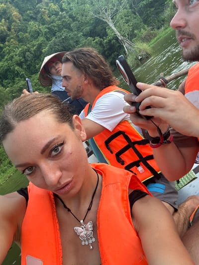 The image captures a group of people on a boat, likely a tourist excursion, in Yên Sơn Ward, Vietnam. In the foreground, a woman is taking a selfie, wearing an orange life jacket and a butterfly pendant. Her hair is pulled back, and she has makeup on, including winged eyeliner. Behind her, a man with long, wavy hair, also wearing an orange life jacket, is holding a smartphone. To the left, a person wearing a conical hat and a light blue headscarf is visible. The background consists of lush green trees and a body of water. The lighting suggests it is daytime.