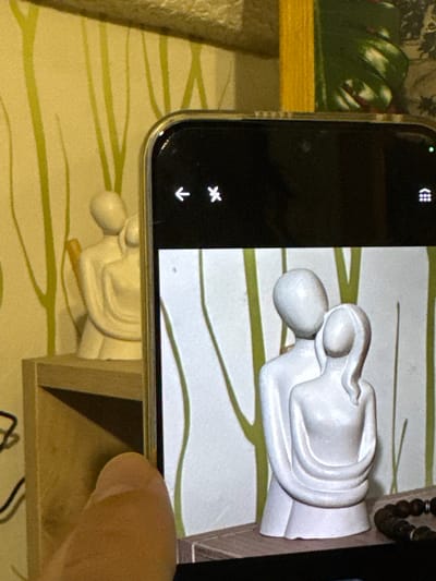 A smartphone is being used to photograph a small, white, reclining statue of two figures embracing, resting on a surface with a green and beige patterned design. The statue is positioned on a wooden shelf, visible underneath the phone. A human finger is touching the screen of the phone. Part of a string of beads and another object with green coloration are visible in the upper left background.