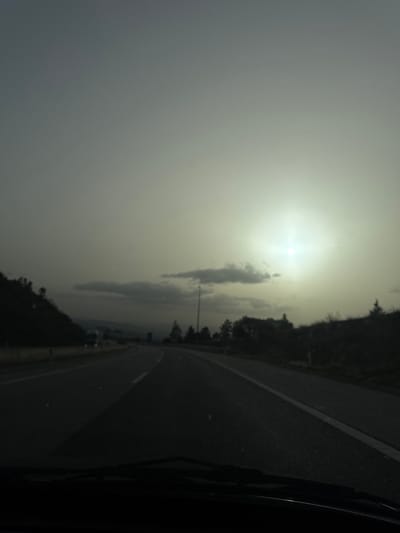The scene depicts a view from inside a vehicle traveling on a highway near Macedo de Cavaleiros, Portugal, under hazy atmospheric conditions consistent with a low sun angle. The upper two-thirds of the frame are dominated by an overcast, grayish-green sky, featuring a bright, diffuse sun in the upper right quadrant with noticeable lens flare. Several dark, elongated clouds are scattered across the mid-sky. The asphalt road, marked with white lines, occupies the lower portion of the image, curving gently to the right into the distance. To the left of the road, a dark, tree-covered hillside is visible, behind which a light-colored truck is partially discernible in the mid-distance. A similar dark, tree-covered embankment runs along the right side of the road, with a tall, thin utility pole positioned between the road and this embankment. The bottom edge of the image displays the top of a vehicle's dashboard and windshield wipers. No individuals are visible.
