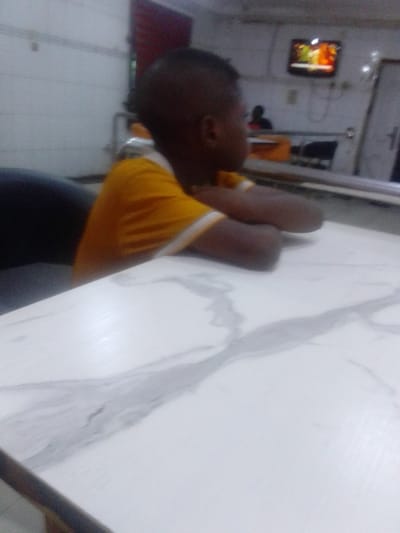 The image shows a young boy in a yellow t-shirt sitting at a white marble table, leaning forward with his arms crossed. The table is in a room with white walls and a black chair behind him. Another person is sitting at the table out of focus in the background. The room appears to be a restaurant or a cafe, and there is a television on the wall in the background playing a program. The coordinates provided locate this scene in Sapele, Nigeria.