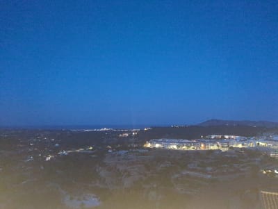 This media file is a wide-angle, high-angle shot of a town at dusk or night, likely in Polop, Spain. The sky is a deep, clear blue, transitioning to a darker hue near the horizon, suggesting the evening or early morning. The town is illuminated by numerous lights, indicating artificial lighting from buildings and streetlights.
The primary subjects are the buildings of the town, which appear to be a mix of residential and commercial structures. A cluster of buildings in the mid-ground stands out, featuring lit windows and external lighting. The landscape surrounding the town is hilly and somewhat rugged, with darker, shadowed areas suggesting vegetation or uneven terrain. In the distance, a body of water, likely the sea, is visible, bordered by a distant coastline dotted with more lights. A mountain range forms the backdrop behind the town.
The scene depicts the town as it comes alive with artificial light as natural light fades. The activity is passive, focused on the existence of the illuminated settlement against the natural landscape. Notable details include the contrast between the dark terrain and the brightly lit town, and the vast expanse of the twilight sky. The overall mood is serene and peaceful, evoking a sense of calm as day transitions to night. There is no visible text in the media file.