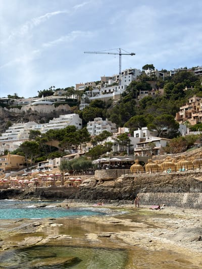 This is a vertical, eye-level, full shot of a rocky cove with a beach resort in Port D'Andratx, Spain. In the foreground, rocky formations are partially submerged in clear turquoise water. In the midground, a beach area has thatched umbrellas and lounge chairs. A stone retaining wall separates the beach from a hillside covered in white apartment buildings and lush green trees. A construction crane is visible in the upper right quadrant of the image, above the buildings. Several people are in the water and on the beach, some sunbathing, some swimming, and one person is standing near the water's edge with another person.