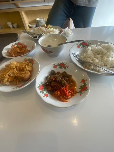 A table setting for one, likely in a casual eatery in Medan, Indonesia, features several small plates of food and a bowl of soup or sauce. The table is white and reflects light. In the foreground, a white, shallow bowl with a floral pattern holds a portion of red chili paste with visible chopped chili peppers and some oil. To the left, another similar bowl contains a fried dish with a crispy, golden-brown batter, possibly a type of fried chicken or seafood. Further left, a smaller oval dish holds what appears to be dried or crispy fried food items. Behind these, a larger white plate holds a generous serving of white rice, with a fork resting on it. A white bowl with a red and green floral pattern contains a pale liquid, possibly soup or a sauce. In the background, a stack of small white plates is visible, along with some decorative cups. A person's legs, clad in blue jeans, are visible in the upper right portion of the image, suggesting they are seated at the table. The overall impression is of a simple meal served in a clean, well-lit environment.