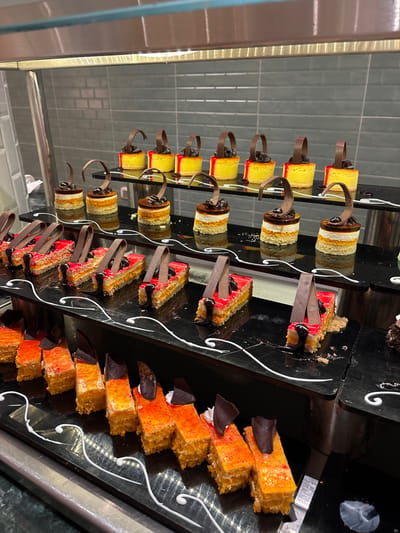 The image shows a display of cakes in a buffet-style setting. The cakes are arranged on multiple tiers. The cakes are decorated with chocolate curls and red and orange frosting. The buffet is likely inside a restaurant or hotel in Al Ahia', Egypt, but the exact location is unknown.