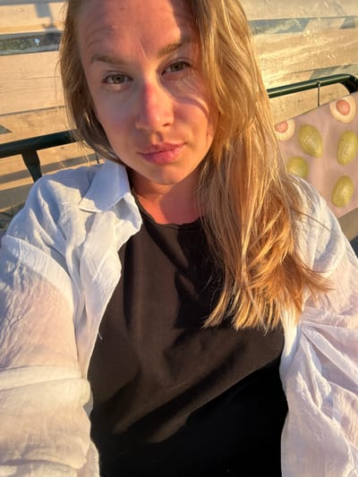 The image is a close-up, selfie-style shot of a woman with blonde hair, wearing a black t-shirt and an unbuttoned white shirt. She is outdoors, with sunlight illuminating her face and hair, casting shadows from a wooden structure behind her. A green metal railing is visible, along with a pink fabric with an avocado print. The environment suggests a relaxed outdoor setting, possibly a garden or a patio. The location is specified as Zamosh'e (Zaklinskoe S/P), Russia.