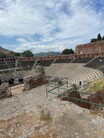 The image shows an outdoor amphitheater in Taormina, Italy, likely the Ancient Theatre of Taormina, with tiered seating made of stone and brick. In the foreground, there are scattered rocks and dry grass. Metal railings with a green finish line some of the walkways and stairs. To the left, there is a stage setup with scaffolding, lighting equipment, and large black cylindrical objects. Several people are visible in the amphitheater, seated in the stands or walking around. The background features rolling hills and mountains under a cloudy blue sky. The architecture suggests historical significance, with remnants of brick walls and arched openings visible along the upper sections of the amphitheater.